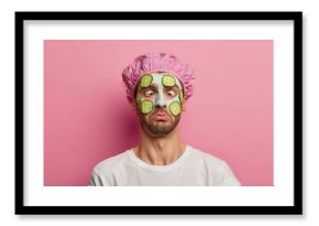 Young man crosses eyes, applies cream mask on face with cucumber, has beauty session, wears bath cap, reduces dark dotes on complexion, isolated over pink background. Facial treatment concept