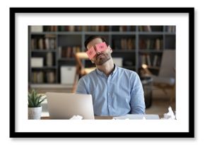 Exhausted tired businessman with painted eyes on stickers, adhesive notes on face sleeping at workplace, sitting at desk with laptop, unproductive lazy young male dozing, working on difficult project