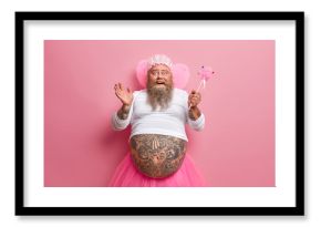 Horizontal shot of joyous funny man with thick beard and big fat belly, plays fairy on costume party, chills during spare time, sends positive vibes, works as animator, isolated over rosy background