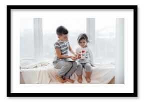 Brother and sister are sitting on the windowsill and reading a book. Happiness, family