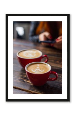 Dwa czerwonej filiżanki kawy na drewnianym tle z latte sztuką. Stolik w kawiarni. Koncepcja łatwego śniadania. Małe i duże ceramiczne kubki. Widok z góry. Dziewczyna z telefonem w tle