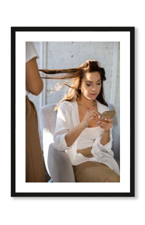 Happy woman hairdresser curl hair of smiling female during chatting social networks use smartphone