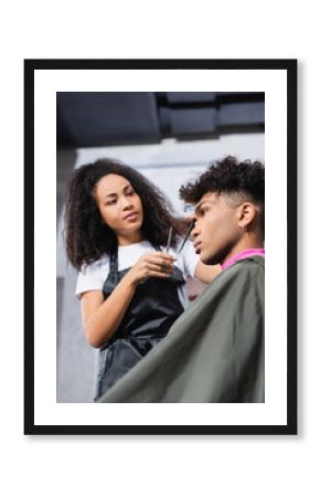 Low angle view of african american client in cape sitting near hairdresser with scissors on blurred background
