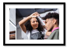 Low angle view of smiling african american hairdresser holding scissors near curly hair of client on blurred foreground