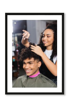 Smiling african american client in cape sitting near hairdresser cutting hair on blurred background