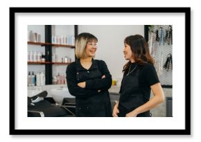 Happy young Asian female hairstylist colleagues working in salon standing wearing apron and spectacles talking to each other