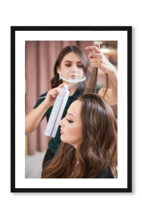 Female hairstylist in protective mask applying hair spray on woman curls. Professional hairdresser fixing client hair locks with hair lacquer while doing hairstyle in beauty salon. Focus on woman.