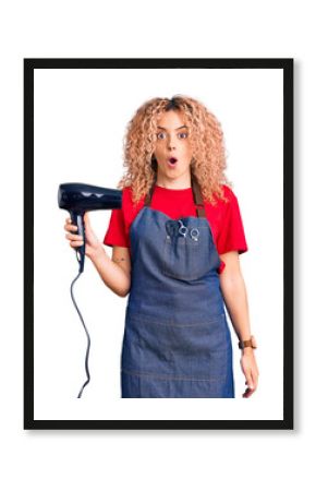 Young blonde woman with curly hair wearing hairdresser apron and holding dryer blow scared and amazed with open mouth for surprise, disbelief face
