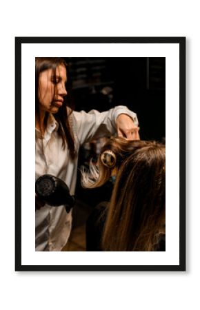 professional woman stylist puts female hair with dryer and round comb in hairdresser.