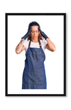 Young hispanic woman with tattoo wearing barber apron and gloves suffering from headache desperate and stressed because pain and migraine. hands on head.