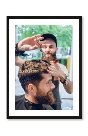 Dedicated male hairstylist using scissors and plastic comb while giving a cool haircut to a redhead bearded young man in a trendy beauty salon