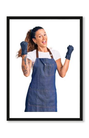 Young hispanic woman with tattoo wearing barber apron and gloves screaming proud, celebrating victory and success very excited with raised arms