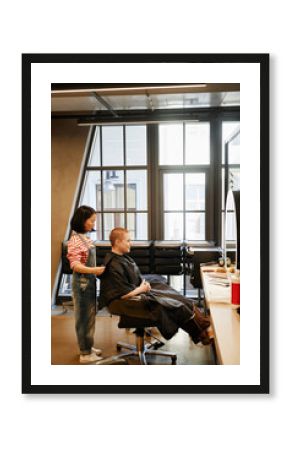 Vertical full length portrait of young woman with buzzcut sitting in beauty salon chair with female hairstylist working