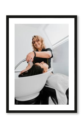 Positive hairstylist woman washing his client's brunette hair. White hairdresser salon interior. Hispanic client in a beauty salon.