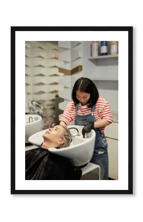Vertical portrait of young hairstylist washing hair of male client n in salon sink and giving professional haircare treatment for dyed hair