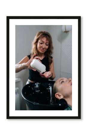 Vertical portrait of hairdresser foaming shampoo on long blonde hair of beautiful female client. Professional hairstylist washing head of young woman in beauty hairdressing salon.