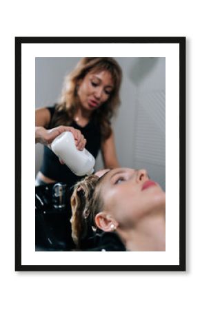 Vertical shot of professional hairstylist foaming shampoo on long blonde hair of beautiful female client. Skilled hairdresser washing head of young woman in beauty hairdressing salon.