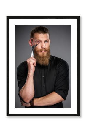 Serious hipster man with beard and mustashes holding scissors looking through it, over grey background
