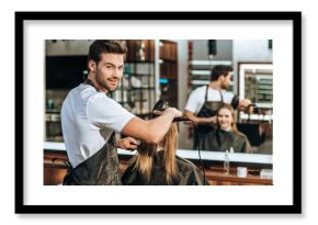handsome young hairstylist smiling at camera while drying hair to beautiful young woman in beauty salon