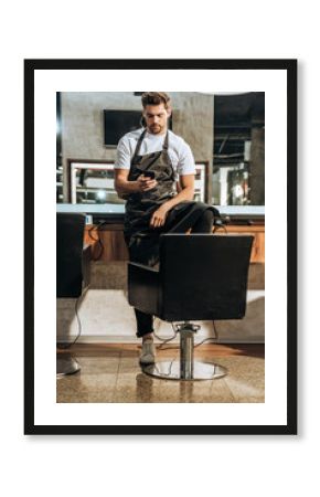 handsome young hairstylist in apron using smartphone in beauty salon
