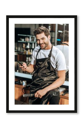 handsome young hairdresser holding smartphone and smiling at camera in beauty salon