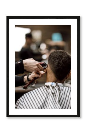 The barber makes a razor cut hair back and sides for a stylish black-haired man sitting in the armchair in a barbershop