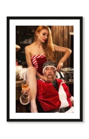 Sexy redhead hairdresser sitting on table and shaving male client in barbershop. Shocked man in santa claus costume drinking beer while pretty woman servicing. Concept of grooming.