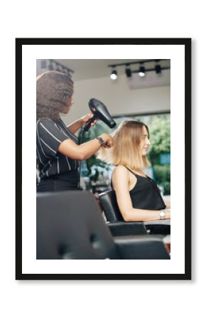 Stylist using professional dryer when blowdrying hair of young pretty woman in her salon