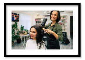 Pure art. Young pretty woman is sitting in the armchair in a beauty salon getting her hair dried by a smiling professional stylist.
