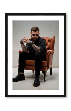 Bearded tattooed man sits on a vintage chair with a thoughtful look