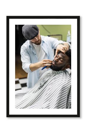 Side view of serious man with stylish modern haircut looking forward in barber shop. Hand of barber keeping straight razor and cutting trendy stripes on head of client. Concept of shaving.