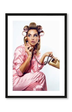 A beautiful young showy girl with bright pin-up make-up speaks on a golden dial telephone in pink striped silk pajamas and pink curlers.