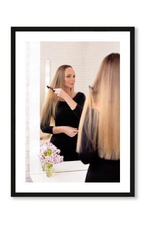Young beautiful woman brushing her long healthy blonde hair and looking at the mirror in hairdressing salon