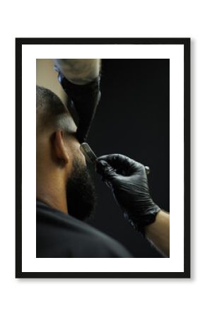 Barber in latex gloves shaving beard of client in barbershop