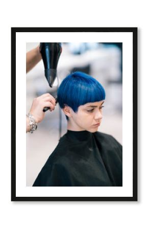 A young woman with vibrant blue hair sits in a salon chair while a hairdresser blow-dries and styles her short haircut. Professional hair care in progress.