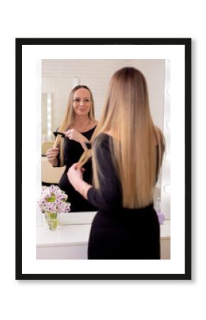 Young beautiful woman brushing her long healthy blonde hair and looking at the mirror in hairdressing salon