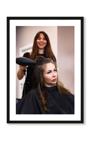 Smiling hairdresser using black hair dryer in salon for chic hairstyle serious brunette woman. Hairstylist lady dried head in barber shop. Customer service at hair salon, haircare. Copy ad text space