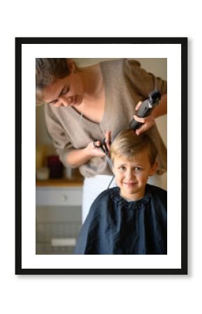 A little blond boy gets a haircut, sitting in a salon chair with a cape. The hairdresser holds scissors, a comb, and a hair clipper. The child is calm and enjoying the experience. 