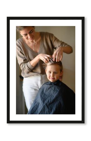 A little blond boy enjoys his new haircut as the hairdresser styles his hair with mousse. He is happy, laughing, and having fun in the salon chair. Concept of childhood grooming and self-care.