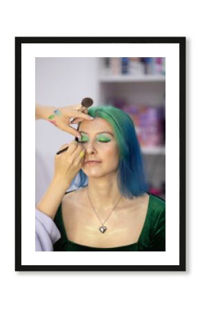 woman with green hair is getting her eyes done by a stylist. The stylist is using a brush to apply makeup to the woman's eyes. The woman is relaxed and comfortable with the process