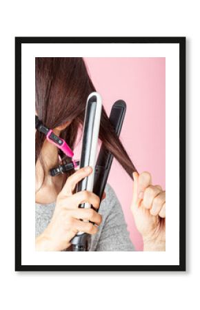 A beautiful young caucasian woman is using sectioning clips and heat hair iron wand for straightening or curling her hair. She is against pink background. DIY beauty concept with electircal tools.