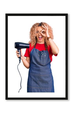 Young blonde woman with curly hair wearing hairdresser apron and holding dryer blow smiling happy doing ok sign with hand on eye looking through fingers