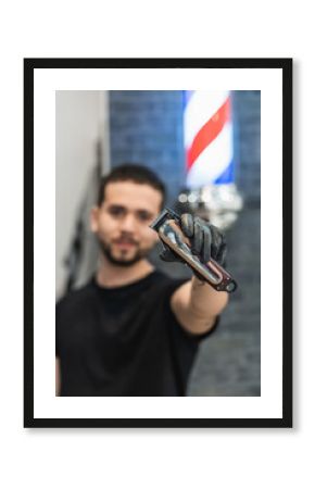Vertical portrait of a barber in his barbershop showing to the camera a hair clipper. Focus on the machine.