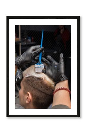 The hands of a girl in black gloves paint the hair of a guy's client white with a brush at work in a barbershop
