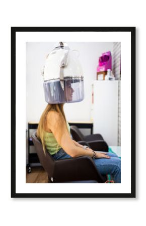 Vertical photo of a pretty woman sitting in a hair salon under a hooded hair dryer. Hair dryer helmet. Dry your hair in a beauty center after a wash.