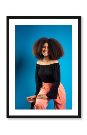 Young woman with black hair, leaning on a wooden stool, posing for a photo.