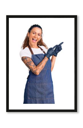 Young hispanic woman with tattoo wearing barber apron and gloves smiling and looking at the camera pointing with two hands and fingers to the side.