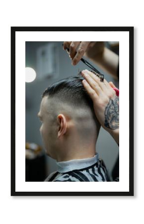 A barber man with a tattoo on his arm makes a haircut to a brunette guy in a barbershop, cuts his hair with a clipper close-up