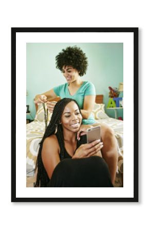 Woman braiding hair of friend holding cell phone in bedroom