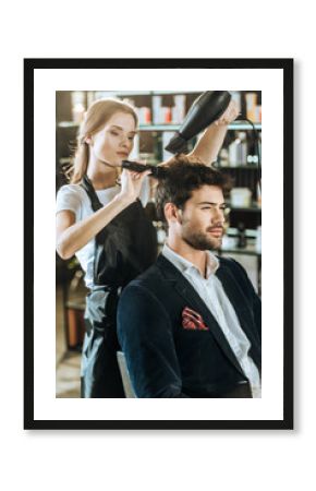 Young hairstylist combing and drying hair to handsome client in beauty salon